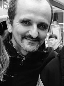 A black and white photo of a white man smiling at the camera, showing just his head and shoulders.