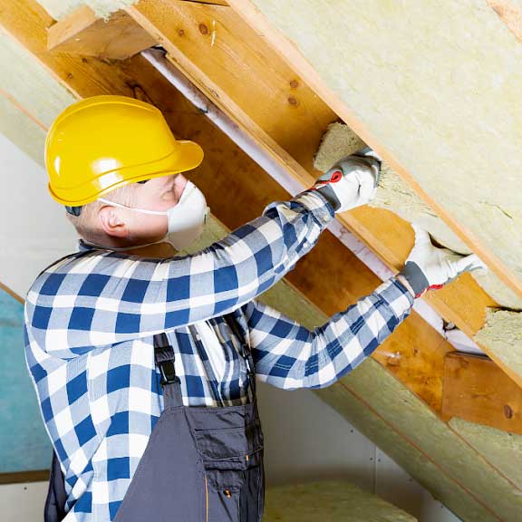 A photo of a white man wearing overalls and a yellow hard hat. He is installing insulation into a slanted roof.