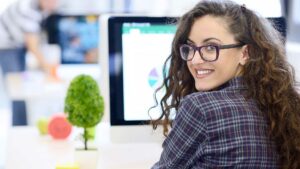 A woman in front of a computer screen, looking back and smiling at the camera.