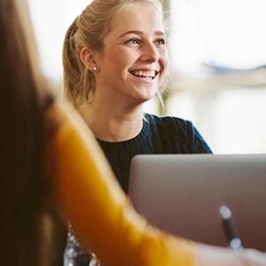 An image of a young white woman sat in front of a laptop, she is smiling at something out of the picture.