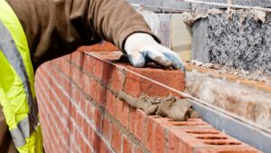 A person wearing a brown jacket and gloves is laying redbricks on a wall.