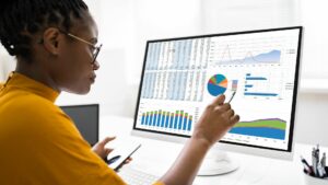 A black woman with glasses, who is wearing a yellow top is looking at a screen with a spreadsheet, data and charts, she is holding a mobile phone and comparing screens. She is entering data on to the main screen.