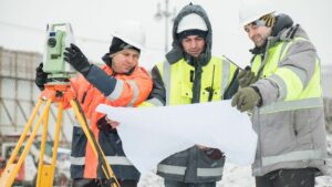 Three men are using an altimeter and looking at plans on a large piece of paper. Ther are all dressed in safety equipment suitable for a building site. They are all wearing high vis warm jackets, one is orange the others are yellow. They all have walky talkies. It is snowing and they are on a construction site.