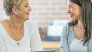 A middle aged white woman with short cropped hair is sat facing a young Asian woman. They are both smiling, and are in conversation. The white woman is holding a tablet computer and is sharing information with her colleague. The white woman is wearing a very light blue long sleeve top, and a gold necklace. The Asian lady is wearing a grey cardigan.