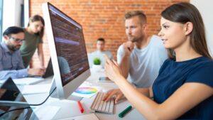 A white woman with long brown hair is pointing at a computer screen with code on it. A white man sat next to here is also looking at the screen, she is teaching him about what is on the screen. They are sat in an office and other colleagues are also working on laptops. They are looking a data analytics