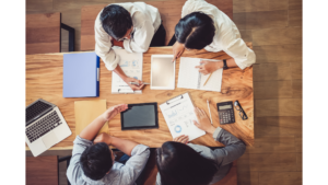 Birds eye view of four people sat at a table looking at tablets and sheets of paper with charts on.