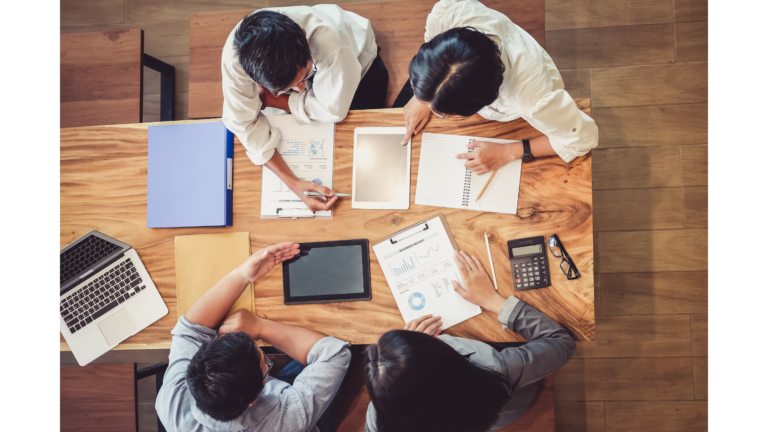 Birds eye view of four people sat at a table looking at tablets and sheets of paper with charts on.