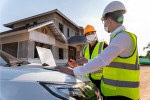 Two men wearing hi-vis jackets, hard hats and face masks are looking at a laptop. In the background is a house being built.