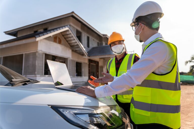 Two men wearing hi-vis jackets, hard hats and face masks are looking at a laptop. In the background is a house being built.