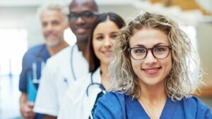 Four people wearing scrubs are stood looking into the camera. The are stood in height order one behind another. The person closest to the camera is a white woman with blond shoulder length curly hair and glasses. She is smiling. She is wearing blue scrubs. There is a lady just behind her with long brown hair, she has white scrubs on and is wearing a stethoscope. There is a tall black man behind her. He has a shaved head. he is wearing white scrubs and a stethoscope. Behind him and very out of focus is a white man with white hair and a beard. He is wearing blue scrubs and a stethoscope and holding a light blue folder.