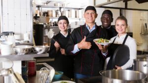 Four people are stood in a catering kitchen there is lots of kitchen equipment in the room including bowls, sauce bottles, whisks and pans. The four people are three chefs and a waitress, they are holding a plat of food in front of them and the person holding the food has their thumb up in front of them. They are all smiling. There are two men and two women.