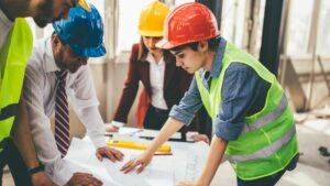 Four people are stood on a building site, they have plans on a table as well as spirit levels and pencils. They are in conversation. There is a woman wearing a high vis tabard, and a red hard hat who is leading the conversations. There is a man in a shirt and tie with a blue helmet on, there is a woman in a yellow helmet and a red blazer with a white shirt. There is a man with a yellow helmet and a high vis tabard over a white t-shirt.