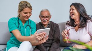 Three people are sitting on a sofa looking at a tablet computer. There is a younger white woman with blonde hair, she is wearing scrubs and there is a stethoscope draped around her shoulders - she is holding the tablet in front of the other two people. An older man with white receding hair is looking at the tablet. He is smiling. He has a check shirt and a brown sweatshirt on. There is also a lady with shoulder length brown hair. She is laughing, and holding her glasses out in front of her. The image is for the skills bootcamp in health and social care.