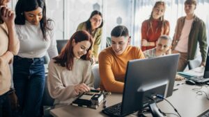 A group of young adults are sitting and standing looking at two people working on a computer motherboard. A young woman with burgundy hair is placing a chip in to the mother board.