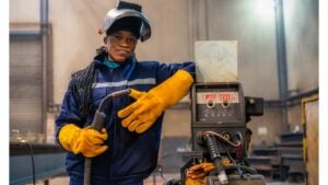 A young black woman with long braided hair, in standing next to a welding rig. She is holding the welder. She is wearing a visor which is open so that you can see her face, she is wearing fire proof overalls and thick gloves. She looks very confident.