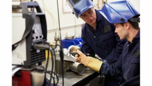 An older white man is teaching a younger white man to weld. They are both wearing safety overalls and safety gauntlets. The young man is holding a welding torch and is preparing to weld a cylindrical shape on to some other metal. They are both wearing blue visors which are open. They are in conversation.