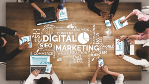 Birds eye view of seven people sat around a large wooden table. They are on their laptops and phones. In the centre of the table is a word map that includes text like digital marketing, S E O, advertising strategy, website promotion