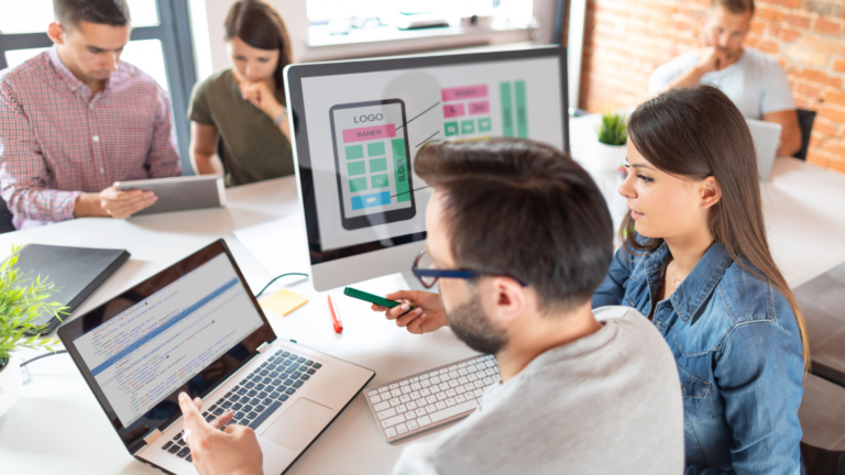 A white woman with long brown hair is pointing at a computer screen with code on it. A white man sat next to here is also looking at the screen, she is teaching him about what is on the screen. They are sat in an office and other colleagues are also working on tablets. They are looking a data analytics