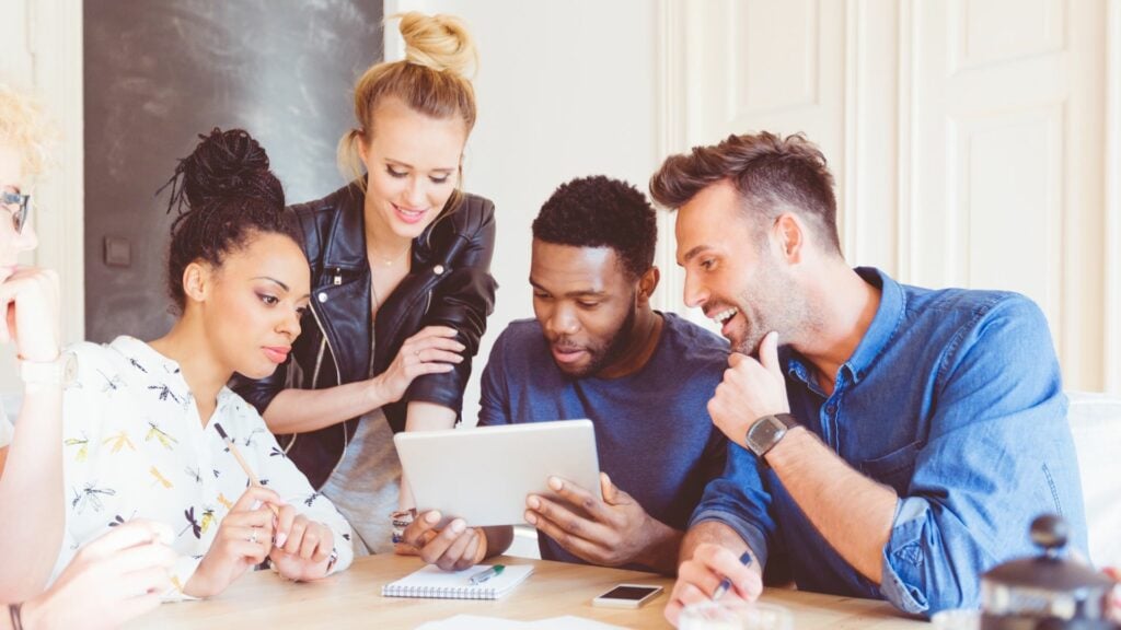 A black man is holding a tablet computer he is showing three other people something on the screen. they all look interested and have a note pad ready to take notes. There are two women, both or who have their hair in buns. One is wearing a leather jacket. There is also a white man holding a pen.