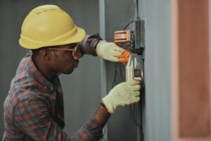 A black man wearing a yellow hard hat and yellow gloves tinkering with a fuse box