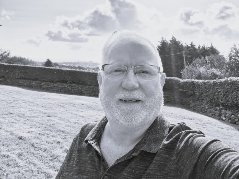 A black and white photo of an older man with a beard taking a selfie with a view of field behind him.