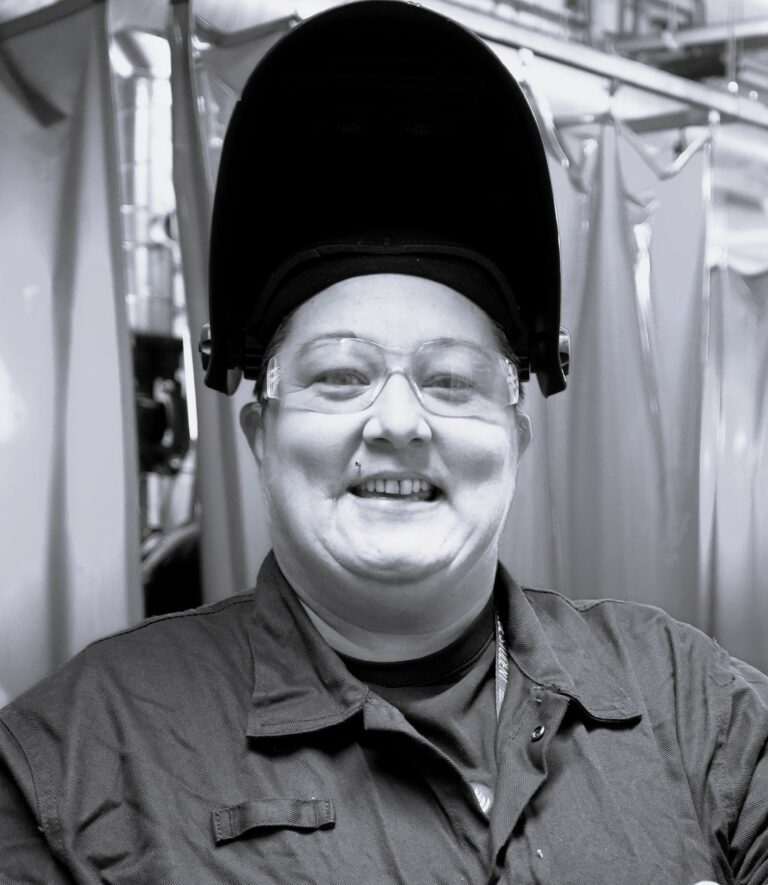 A black and white photo of a smiling woman wearing welding protective gear