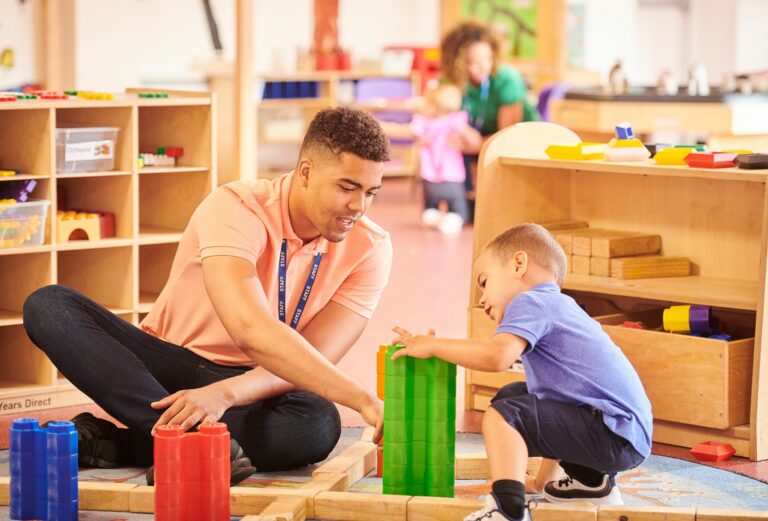 A man sat on the floor in a nursery with a little boy playing with building bricks