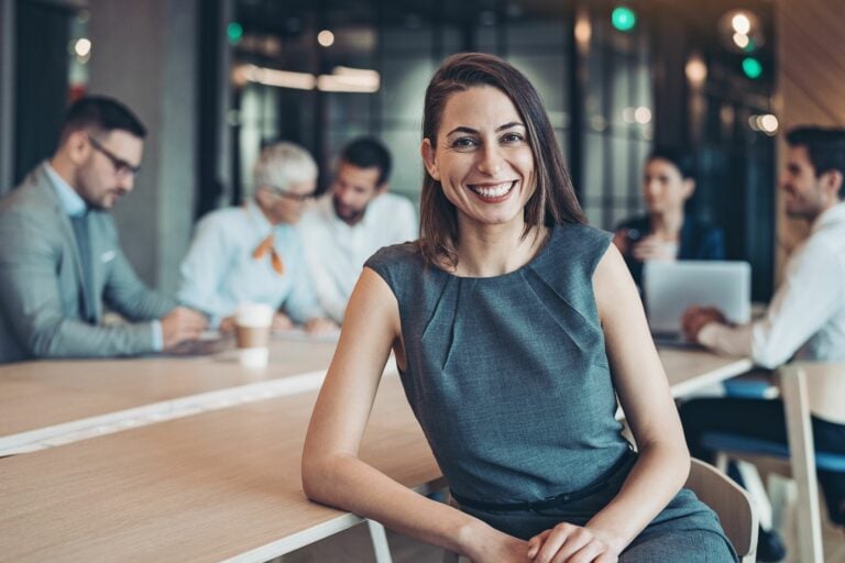 A woman smiling at the camera. In the background there is a team of workers sat around a big table.