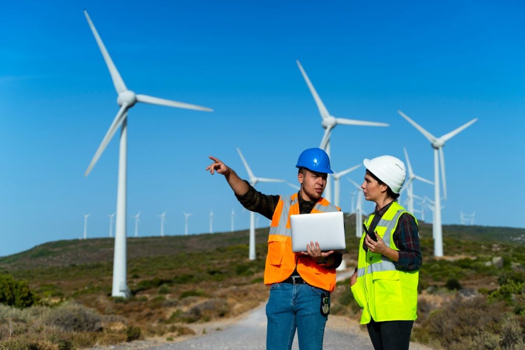 Two people wearing hi vi vests and hard hats surrounded by wind turbines